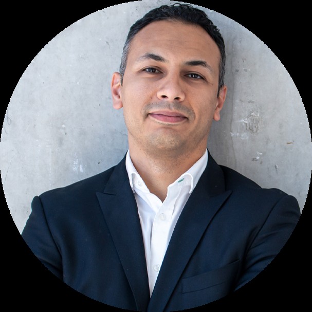 Man with short dark hair wearing a dark navy blazer over a white shirt, smiling against a grey concrete wall, framed in a circular crop.