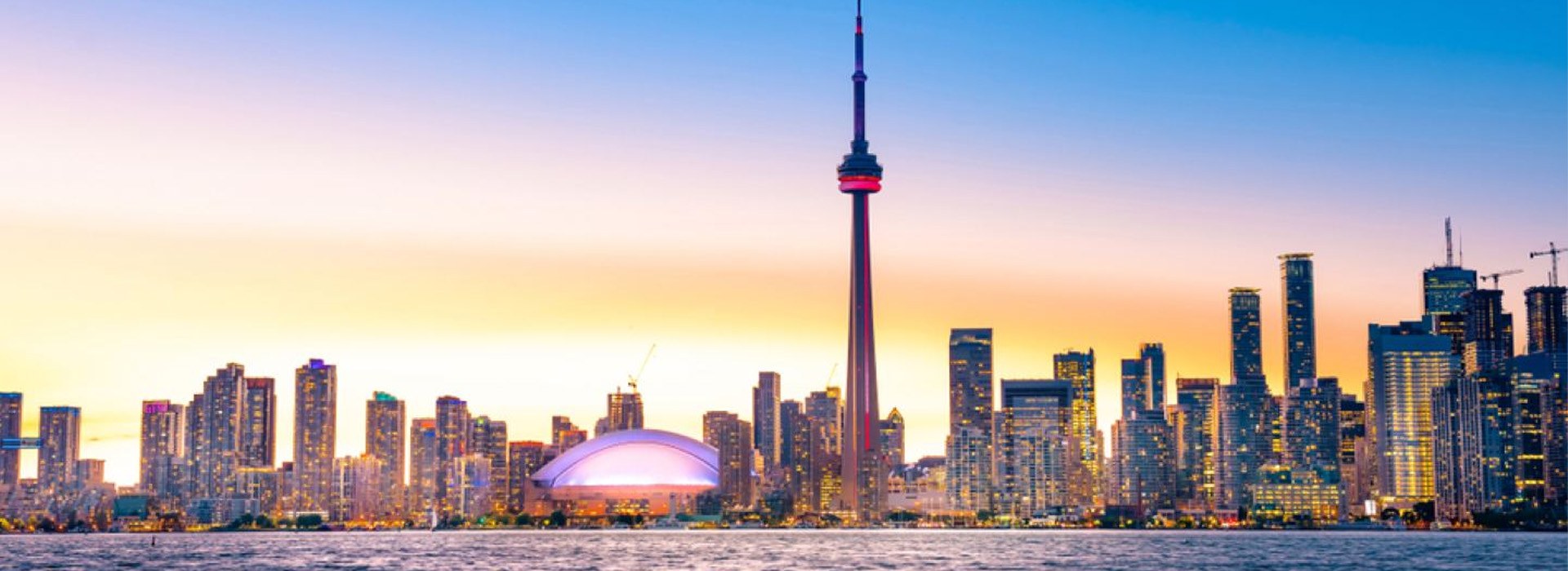 Toronto skyline at sunset with the CN Tower, illuminated Rogers Centre dome, and waterfront skyscrapers glowing against an orange and blue sky.