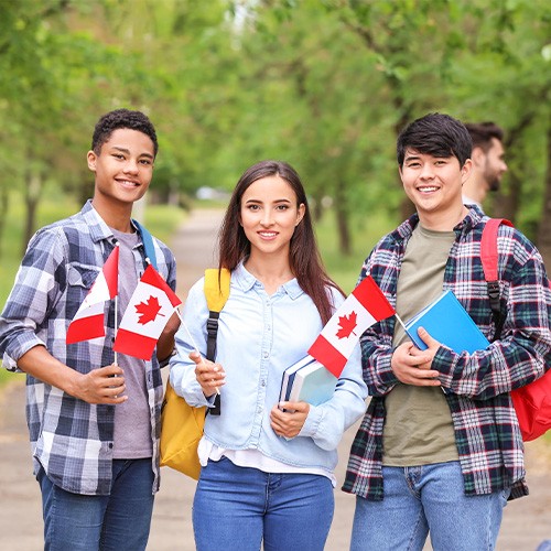 Three young adults stand outdoors holding Canadian flags and books, smiling at the camera.